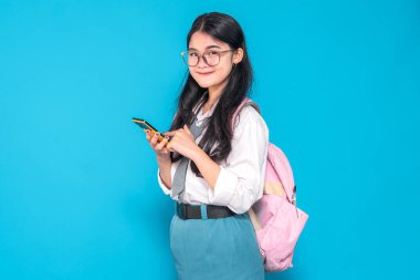 Endonezyalı bir lise öğrencisi gözlüklü ve sırt çantalı, akıllı telefonunu kullanırken gülümsüyor. Meşgul ve mutlu görünüyor, parlak mavi bir arkaplanda duruyor..