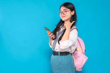 Endonezyalı bir lise öğrencisi, gözlüklü ve sırt çantalı, akıllı bir telefonu tutarken güvenle gülümseyen ve parlak mavi bir arka plana karşı olumlu bir hareket yapan.