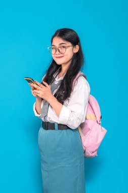 Endonezyalı bir lise öğrencisi gözlüklü ve sırt çantalı, akıllı telefonunu kullanırken gülümsüyor. Meşgul ve mutlu görünüyor, parlak mavi bir arkaplanda duruyor..