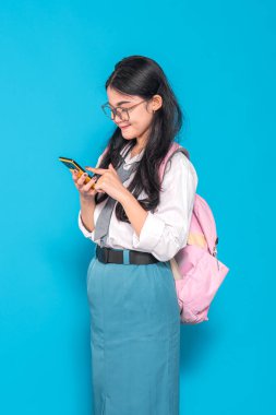 Endonezyalı bir lise öğrencisi gözlüklü ve sırt çantalı, akıllı telefonunu kullanırken gülümsüyor. Meşgul ve mutlu görünüyor, parlak mavi bir arkaplanda duruyor..