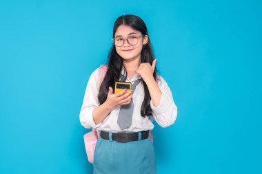 Endonezyalı bir lise öğrencisi, gözlüklü ve sırt çantalı, akıllı bir telefonu tutarken güvenle gülümseyen ve parlak mavi bir arka plana karşı olumlu bir hareket yapan.