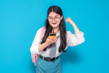 Endonezyalı bir lise öğrencisi, gözlüklü ve sırt çantalı, akıllı bir telefonu tutarken heyecanla gülümseyen ve kollarını zafer pozu vererek parlak mavi bir arka plana karşı koyuyor..