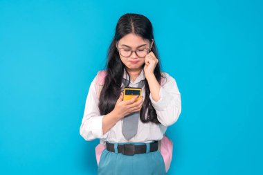 Endonezyalı bir lise öğrencisi gözlüklü ve sırt çantalı, akıllı telefonuna bakarken hayal kırıklığına uğramış görünüyor. Parlak mavi bir arka plana karşı hayal kırıklığına uğramış bir ifade sergiliyor..