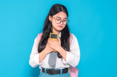 Endonezyalı bir lise öğrencisi gözlüklü ve sırt çantalı duygusal bir ifadeyle akıllı telefonunu göğsüne yakın tutuyor. Parlak mavi bir arkaplan karşısında duygusal görünüyor..