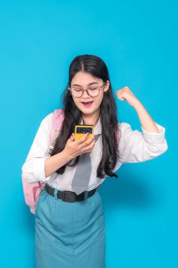 Endonezyalı bir lise öğrencisi, gözlüklü ve sırt çantalı, akıllı bir telefonu tutarken heyecanla gülümseyen ve kollarını zafer pozu vererek parlak mavi bir arka plana karşı koyuyor..