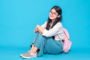 Endonezyalı bir lise öğrencisi gözlüklü ve sırt çantalı rahat bir gülümsemeyle öylece yerde oturuyor. Parlak mavi bir arka planda rahat ve neşeli görünüyor..