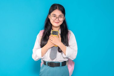 Endonezyalı bir lise öğrencisi gözlüklü ve sırt çantalı duygusal bir ifadeyle akıllı telefonunu göğsüne yakın tutuyor. Parlak mavi bir arkaplan karşısında duygusal görünüyor..