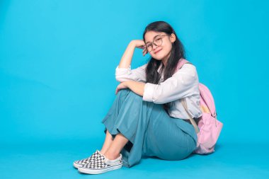 Endonezyalı bir lise öğrencisi gözlüklü ve sırt çantalı rahat bir gülümsemeyle öylece yerde oturuyor. Parlak mavi bir arka planda rahat ve neşeli görünüyor..