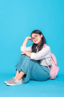 Endonezyalı bir lise öğrencisi gözlüklü ve sırt çantalı rahat bir gülümsemeyle öylece yerde oturuyor. Parlak mavi bir arka planda rahat ve neşeli görünüyor..