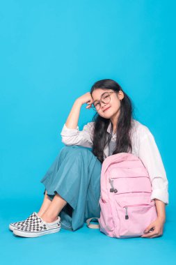 Endonezyalı bir lise öğrencisi, gözlüklü, yerde oturmuş, rahat bir gülümsemeyle başını elinin üzerine koyuyor. Pembe bir sırt çantası onun yanında, parlak mavi bir arka planda..