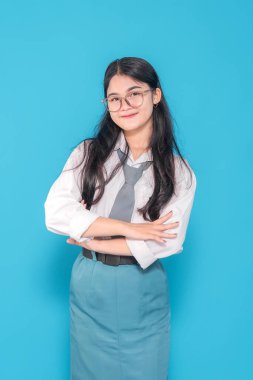 Gözlüklü Endonezyalı bir lise öğrencisi, kollarını kavuşturmuş ve hafifçe gülümsüyor. Parlak mavi bir geçmişe karşı zeka ve kendine güven yayıyor..