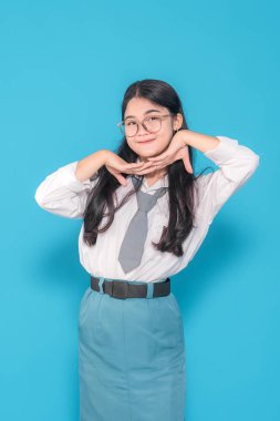 Endonezyalı bir lise öğrencisi gözlüklü, iki eli çenesinin altında şakacı bir şekilde poz veriyor, tatlı tatlı gülümsüyor. Parlak mavi bir arka plana neşe ve cazibe saçar..