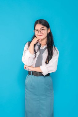 Endonezyalı bir lise öğrencisi gözlüklü, hafif bir gülümseme ve parlak mavi bir arka plana karşı düşünceli bir el pençe hareketi ile özgüvenle poz veriyor..