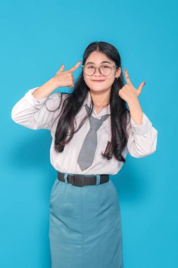 Endonezyalı bir lise öğrencisi gözlüklü, şakaklarını göstererek güvenle gülümsüyor. Parlak mavi bir arkaplan karşısında zekasını ve oyunculuğunu gösteriyor..