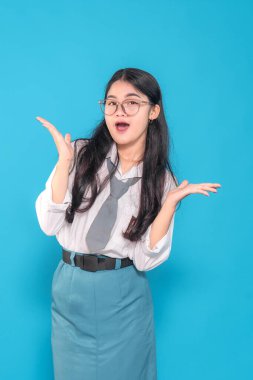 Endonezyalı bir lise öğrencisi okul üniforması ve gözlük takıyor, ağzı açık bir şekilde sürprizi ifade ediyor ve ellerini kaldırıyor. O, parlak mavi bir arka plana karşı meraklı ve heyecanlı görünüyor..