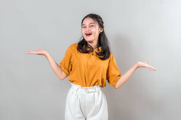Happy young Asian woman with cheerful smile and joyful facial expression, raising both hands in a shrugging gesture, ideal for advertising concepts about choices or fun situations.