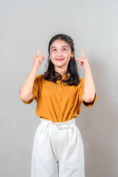 Portrait of a cheerful young woman in a mustard shirt and white pants, smiling while pointing upwards with both hands and looking up, expressing excitement and inspiration.