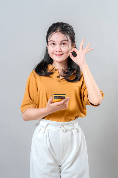 Cheerful young Asian woman smiling with confident expression while making an OK hand gesture and holding a smartphone, representing satisfaction, approval, and positive communication.