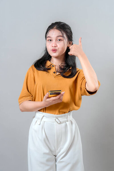 Young Asian woman making a playful kissy face and giving a thumbs up while holding a smartphone. Her expressive face and confident pose convey positivity, approval, and friendliness.