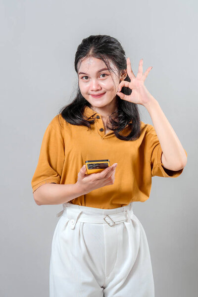Cheerful young Asian woman smiling with confident expression while making an OK hand gesture and holding a smartphone, representing satisfaction, approval, and positive communication.