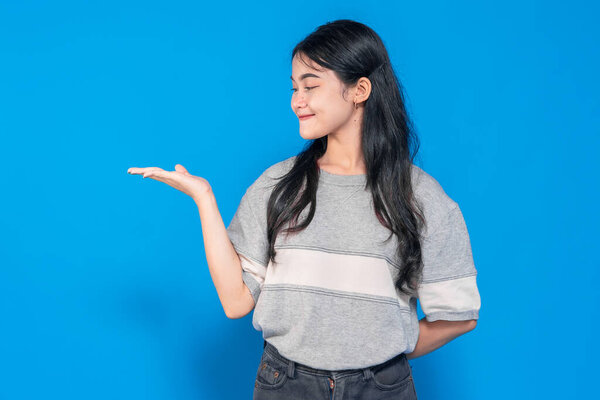 Young Asian woman with a cheerful smile, raising one hand in a presenting gesture. She looks confident and friendly, wearing a casual outfit against a bright blue background.