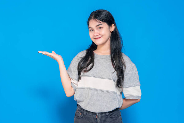 Young Asian woman with a cheerful smile, raising one hand in a presenting gesture. She looks confident and friendly, wearing a casual outfit against a bright blue background.