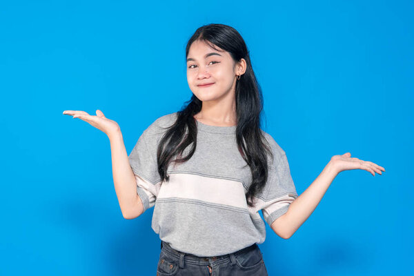 Young Asian woman with a playful smile, raising both hands in a questioning gesture. She looks confident and friendly, wearing a casual outfit against a bright blue background