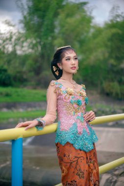 Zarif bir kebaya ve batik giyen Endonezyalı güzel bir kadın nehir kenarında zarif bir şekilde duruyor, doğal bir ortamda duruş, kültürel gurur ve zamansız zarafeti ifade ediyor..