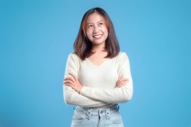 Düşünceli Endonezyalı bir kadın, temiz mavi bir arkaplana karşı kollarını kavuşturarak gülümseyerek küresel iş içeriğine özgüvenli, yaklaşılabilir ve modern bir görünüm sunuyor..