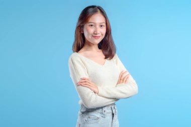 Kendine güvenen Endonezyalı bir kadın, kollarını kavuşturmuş, açık renk bir kazak ve kot pantolon giyiyor, sakince gülümsüyor, modern ve profesyonel bir görünüm sergiliyor..