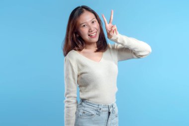 Endonezyalı bir kadın mutlu bir şekilde gülümserken yüzünün yanında net bir barış işareti yapıyor, küresel reklam için uygun temiz mavi bir arka plana karşı neşeli, dostane ve enerjik bir ruh hali sergiliyor..