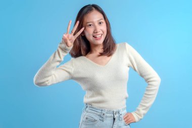 Endonezyalı bir kadın parlak bir şekilde gülümserken, temiz mavi bir arkaplana karşı coşkuyu, dostluğu ve pozitif enerjiyi ifade eden, üç parmak hareketini göstermek için elini kaldırıyor.