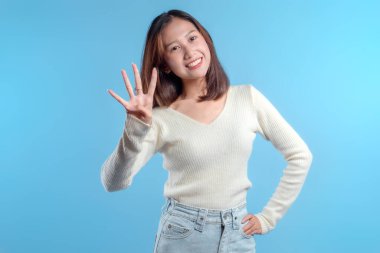 Endonezyalı bir kadın parlak bir şekilde gülümserken dört parmaklı açık bir jest yapmak için elini kaldırıyor, pozitif, berraklık ve dostça enerjisini reklamcılık için uygun temiz mavi bir arka plana karşı ifade ediyor..
