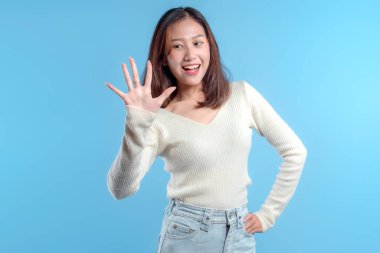 Endonezyalı bir kadın dostça bir jest yaparak elini kaldırırken sıcak bir şekilde gülümsüyor, açık mavi arka planda açık renk bir kazak ve kot pantolon giyiyor..