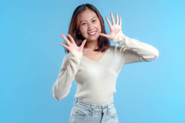Endonezyalı bir kadın parlak bir şekilde gülümserken açık parmaklarıyla iki elini kaldırıyor, açık renk bir süveter ve kot pantolon giyiyor ve temiz mavi bir arka plana karşı pozitif, dostane bir poz veriyor..
