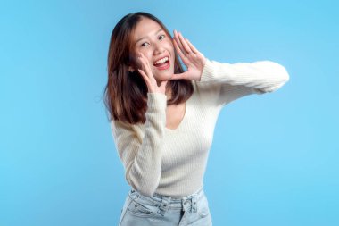 Neşeli Endonezyalı bir kadın iki eli ağzının yanında canlı bir jest yapıyor, parlak mavi bir arka plana karşı enerji, iletişim ve pozitiflik ifade ediyor..