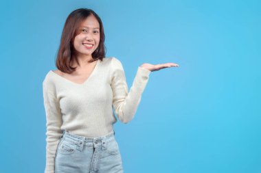 Endonezyalı bir kadın, parlak bir şekilde gülümserken elini yana kaldırıyor, mavi arka planda krem bir süveter ve kot pantolon giyiyor, samimi ve kendinden emin bir şekilde sunum yapıyor..