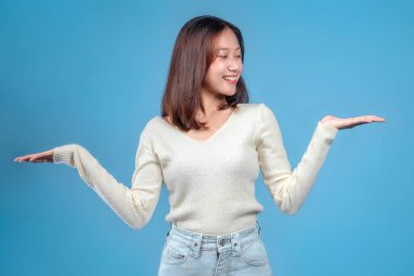 Endonezyalı bir kadın gülümserken iki elini de dengeli bir şekilde kaldırıyor. Mavi arka planda krem bir kazak ve kot pantolon giyiyor. Arkadaşça ve kendinden emin bir tavır sergiliyor..