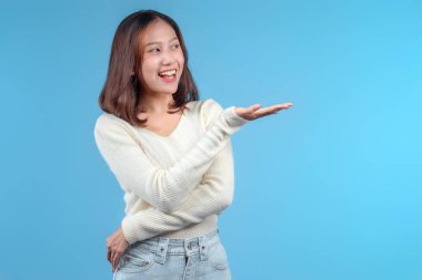 Endonezyalı bir kadın parlak bir şekilde gülümserken elini yana doğru uzatıyor, krem rengi bir süveter ve kot pantolon giyiyor mavi arka planda, samimi ve kendinden emin bir görünüm sergiliyor..