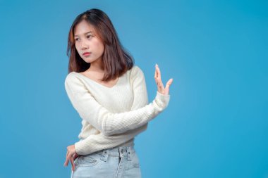 Tedbirli, reddedici bir yüz ifadesi olan Endonezyalı bir kadın elini kenara kaldırıyor ve mavi arka planda krem rengi bir kazak ve kot pantolon giyiyor..