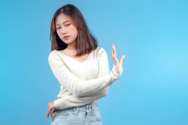 Tereddüt eden, reddeden bir Endonezyalı kadın elini hafifçe yana kaldırıyor, temiz mavi arka planda krem rengi bir kazak ve kot pantolon giyiyor..