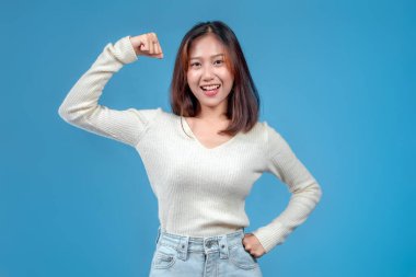 Endonezyalı bir kadın güçlü bir hareketle kolunu kaldırırken kendine güvenle gülümsüyor, temiz mavi bir arka planda krem bir kazak ve kot pantolon giyiyor..