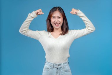 Endonezyalı bir kadın parlak bir şekilde gülümserken her iki kolunu güçlü bir duruşla esnetiyor, temiz mavi arka planda krem bir kazak ve kot pantolon giyiyor..