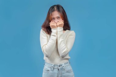 Utangaç, neşeli bir yüz ifadesi olan Endonezyalı bir kadın, yüzünün bir kısmını kaplayan eller, mavi arka planda krem bir süveter ve kot pantolon giyiyor, cazibesini ve gençlik sıcaklığını aktarıyor..