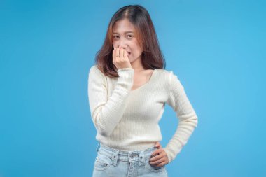 Endonezyalı bir kadın gergin, neşeli bir ifadeyle, parmağını hafifçe ısırırken, temiz mavi bir arka planda, krem rengi bir kazak ve kotla güvenle poz veriyor..