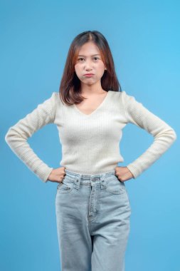 Endonezyalı bir kadın, elleri kalçasında ve kararlı bir ifadeyle, temiz mavi arka planda krem bir kazak ve açık renk kot pantolon giyiyor..