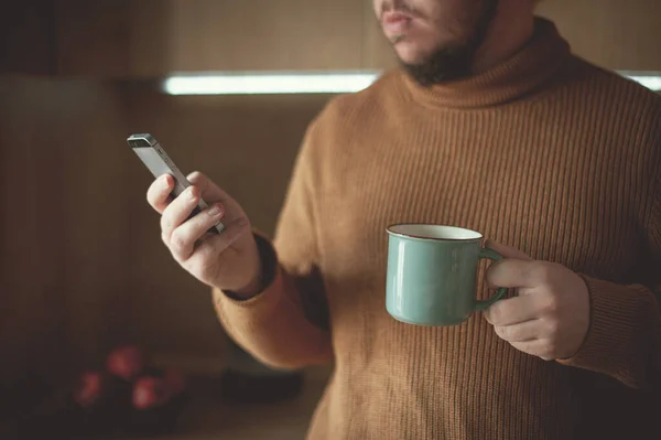 Young man drinks coffee and looks into a smartphone in the kitchen