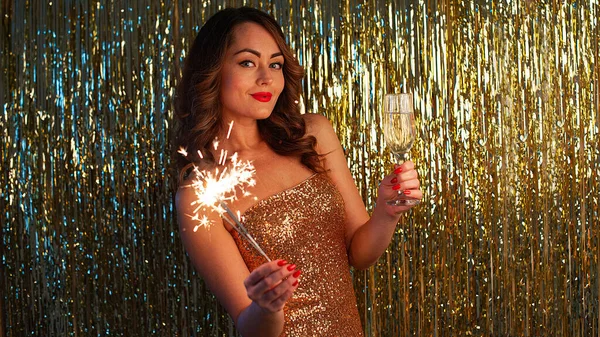 Giovane donna beve champagne e tenendo una scintilla su uno sfondo oro brillante, messa a fuoco selettiva — Foto Stock
