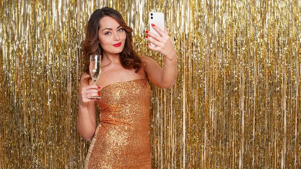Giovane donna beve champagne, fa un selfie su uno smartphone, divertirsi su uno sfondo oro lucido — Foto Stock