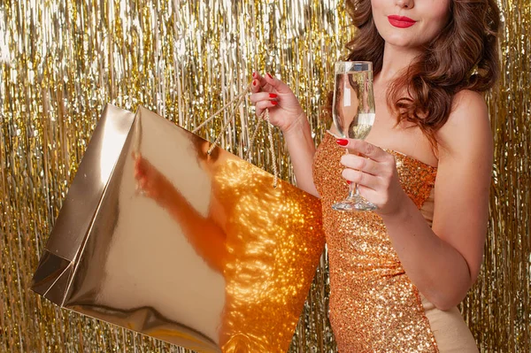 Vrouw in mode kleding met een gouden papieren zak drinkt champagne op een glanzende gouden achtergrond — Stockfoto